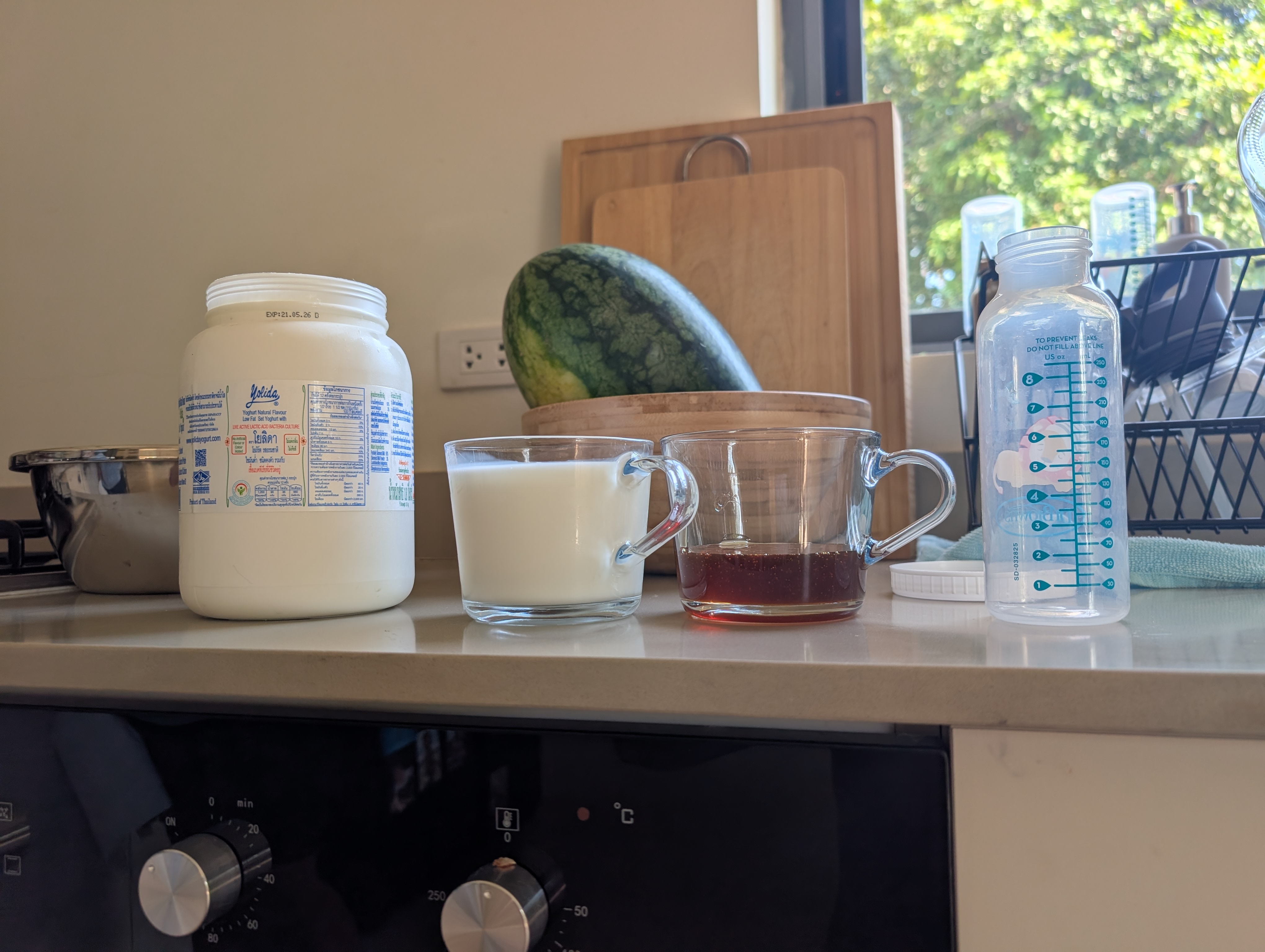 Yogurt batch ingredients on the counter: jar of Yolida yogurt, cup of milk, cup of honey, empty Liv bottle showing the 7oz line